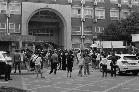A grainy black and white photo of parents standing around outside a school gate in Beijing. They stand facing the school gates, their backs to the camera, waiting. Developed in Ilfosol 3 (1+9).