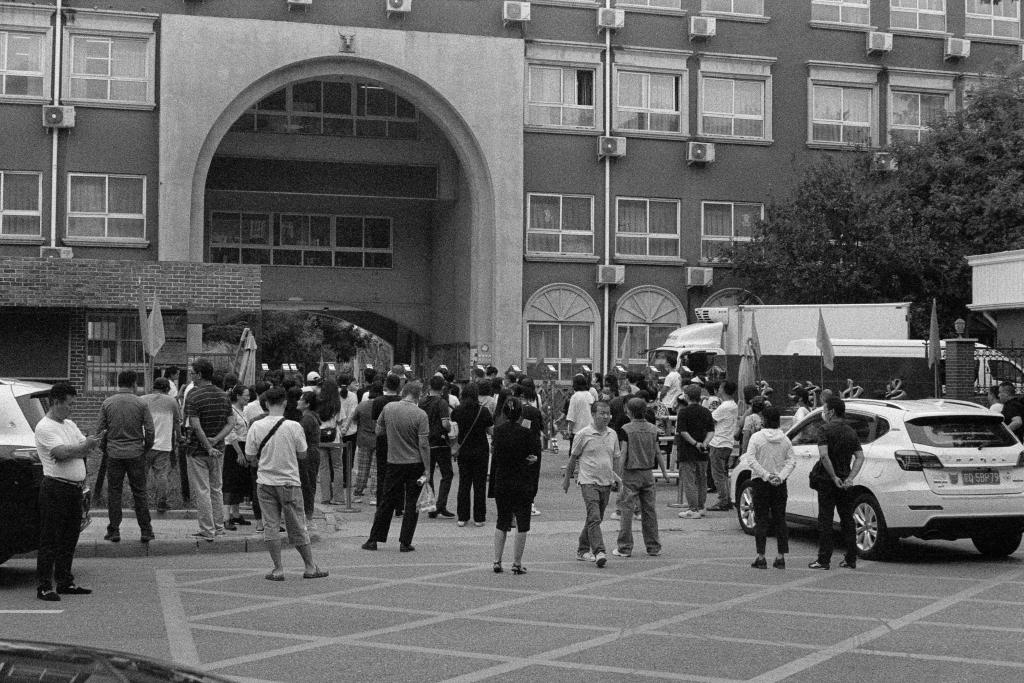 A grainy black and white photo of parents standing around outside a school gate in Beijing. They stand facing the school gates, their backs to the camera, waiting. Developed in Ilfosol 3 (1+9).