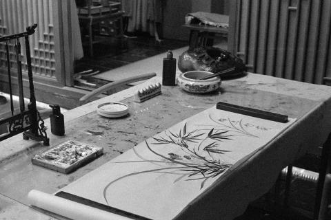 A black and white photo of a writing desk. On the desk is an unfinished ink painting on a scroll of traditional Chinese paper. The brushes hang vertically in a brush holder, the tubes of ink scattered in a small box on the desk. The painting shows a flower, leaves similar to bamboo. On the right of the painting are four Chinese characters. The image is a scan of a negative developed at home in Ilfosol3 (1+9).