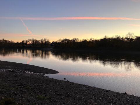 This image was taken on the North bank of the Thames in West London. It looks South across the river. The tide is out and the pebbled bank of the river runs down to the waters edge. Trees line the opposite bank silhouetted against the evening sky. Clouds in the sky are pink with the final rays of the sun.