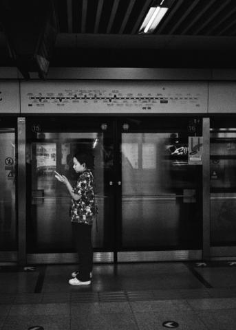 This image was captured at 高米店南 subway station in Beijing. A person stands in front of the platform doors looking towards the left of the image. They are holding a phone. Above the doors is a subway map. The glass of the platform wall results in a layerd image showing reflections from behind the camera and the light from advertisements mounted on the wall in front.