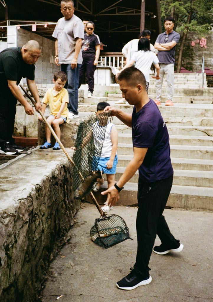 One man holds out a net containing a fish he's just caught. Another prepares to remove the fish from the net. Two children sit watching from the side of the fish tank.